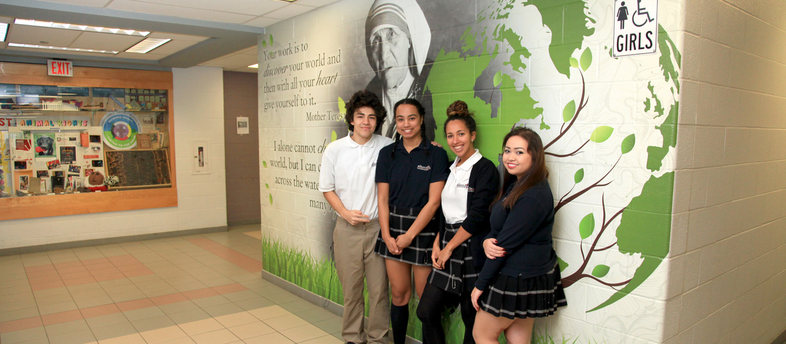 Four students standing in front of a mural depicting a quote and picture of Mother Teresa.
