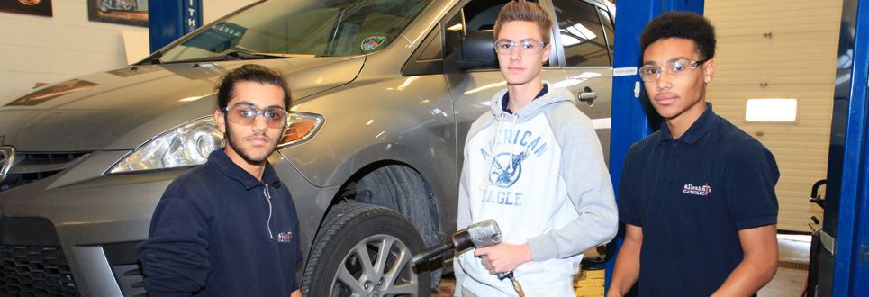 Three male students in an auto shop classroom with car behind