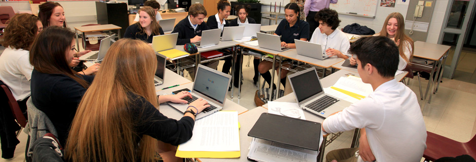 A group of students sitting in a square of desks talking among each other.